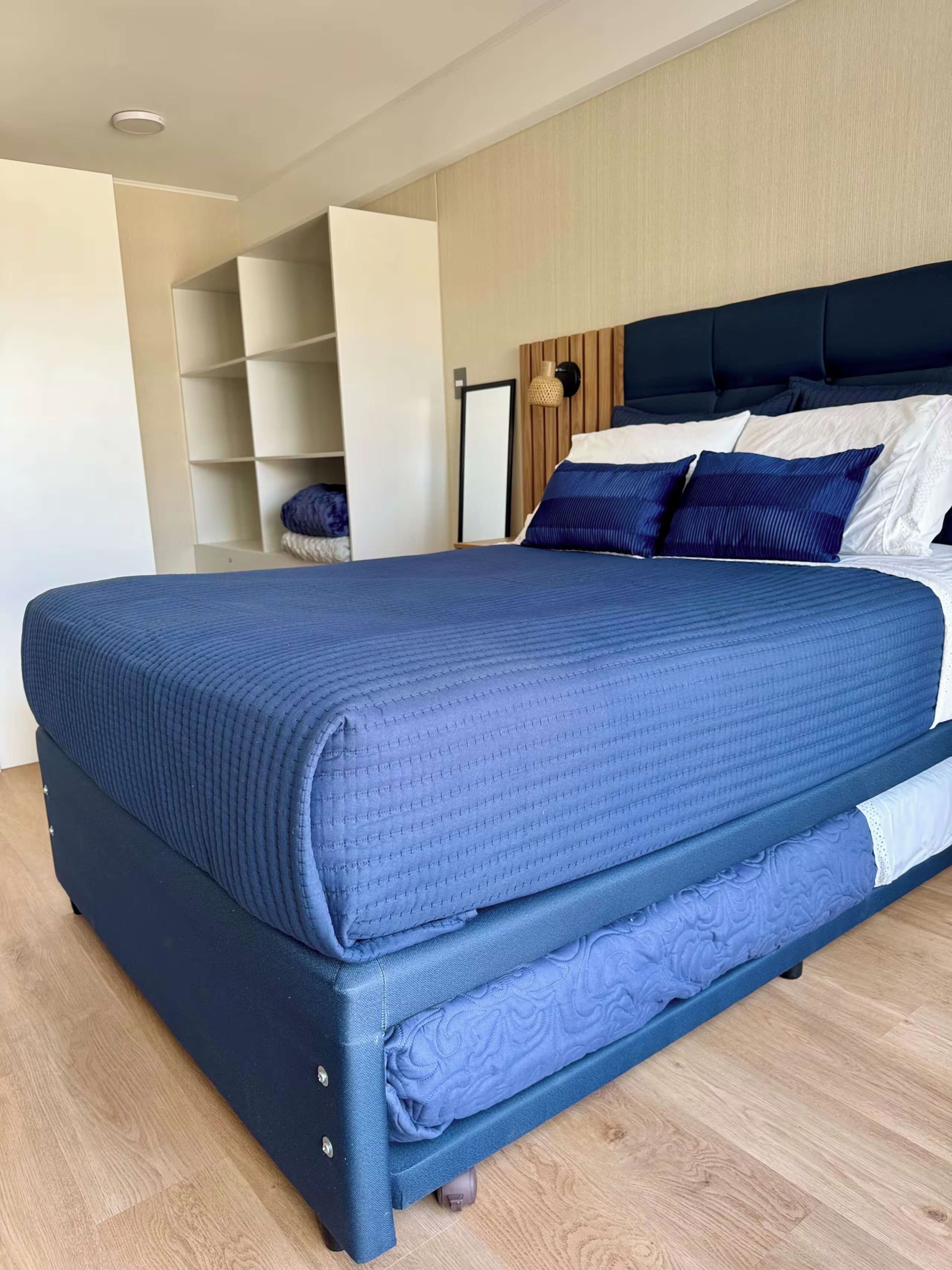 A large bed with a dark blue mattress and matching headboard, a wooden floor, and a built-in shelving unit to the left.