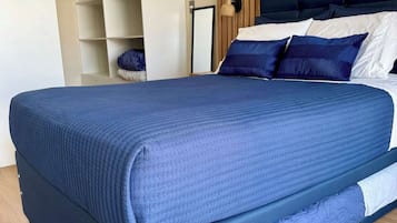 A large bed with a dark blue mattress and matching headboard, a wooden floor, and a built-in shelving unit to the left.