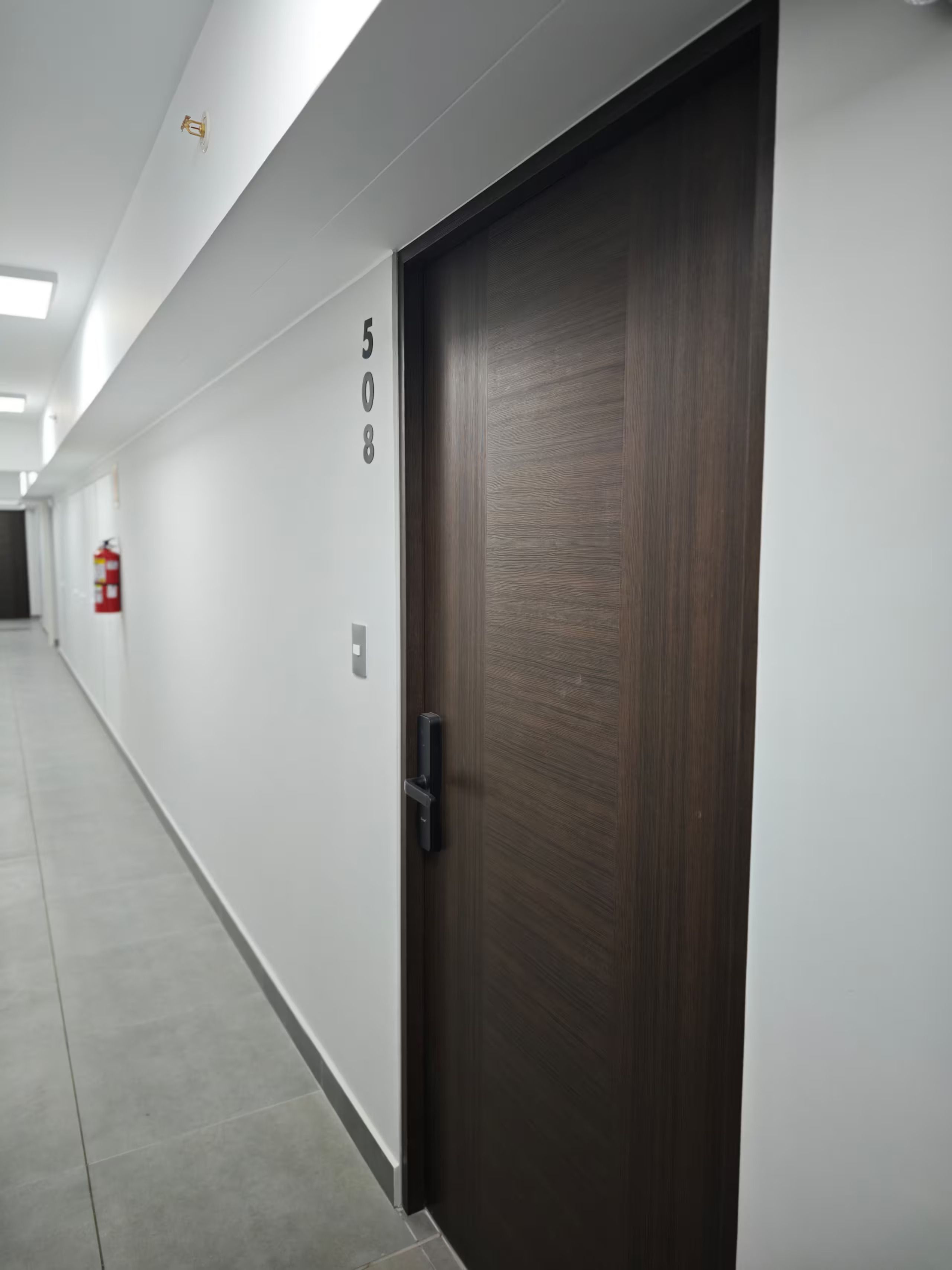 Un pasillo con una puerta de madera oscura, paredes blancas y un extintor instalado en la pared.
