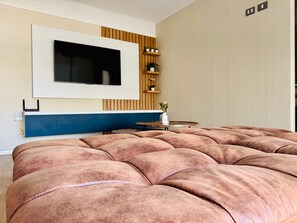 Un dormitorio moderno con una cama grande, un televisor montado, una mesita pequeña y una planta.