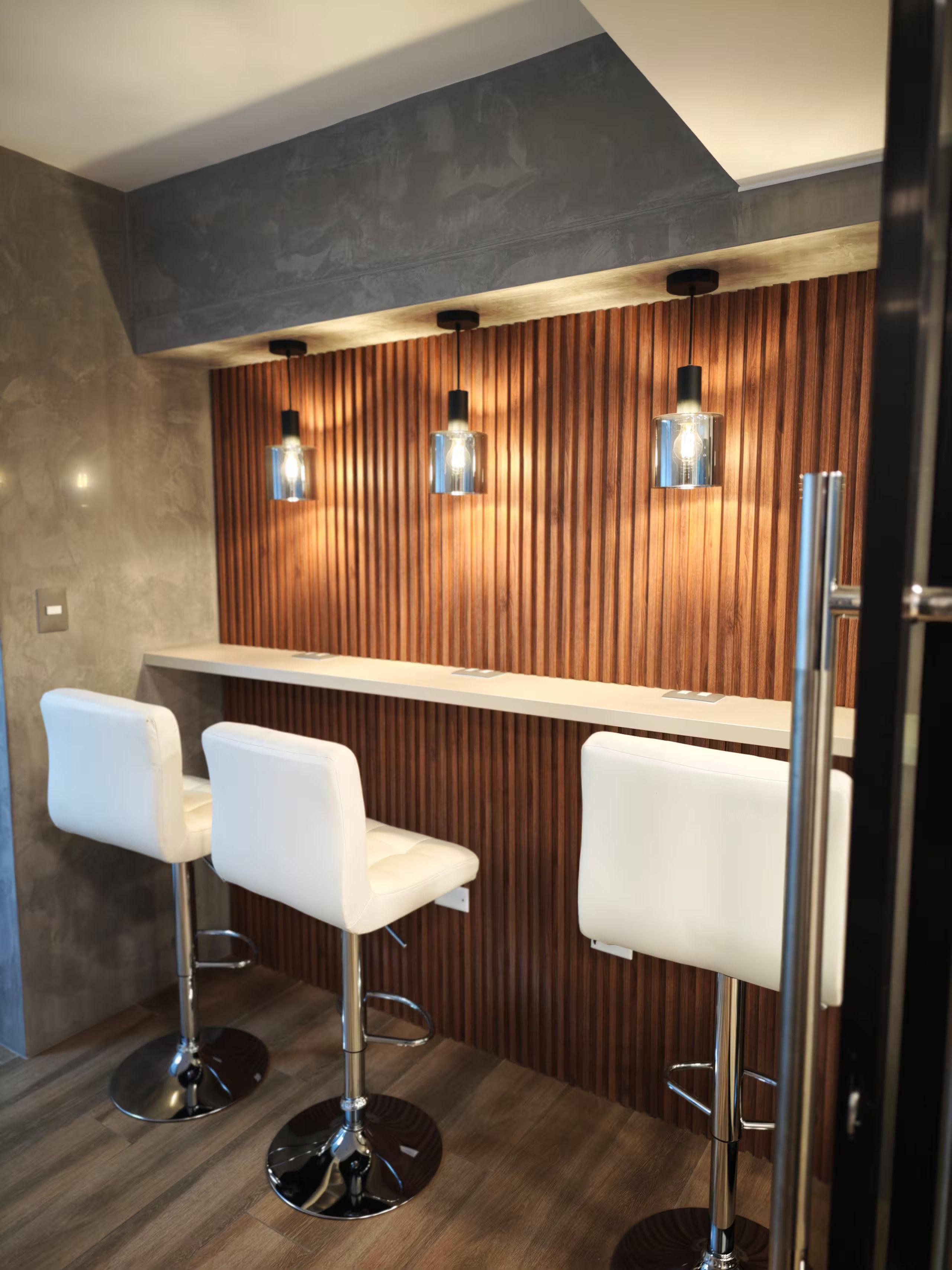 Un espace bar avec trois tabourets de bar blancs, un mur en lambris de bois et des luminaires suspendus.