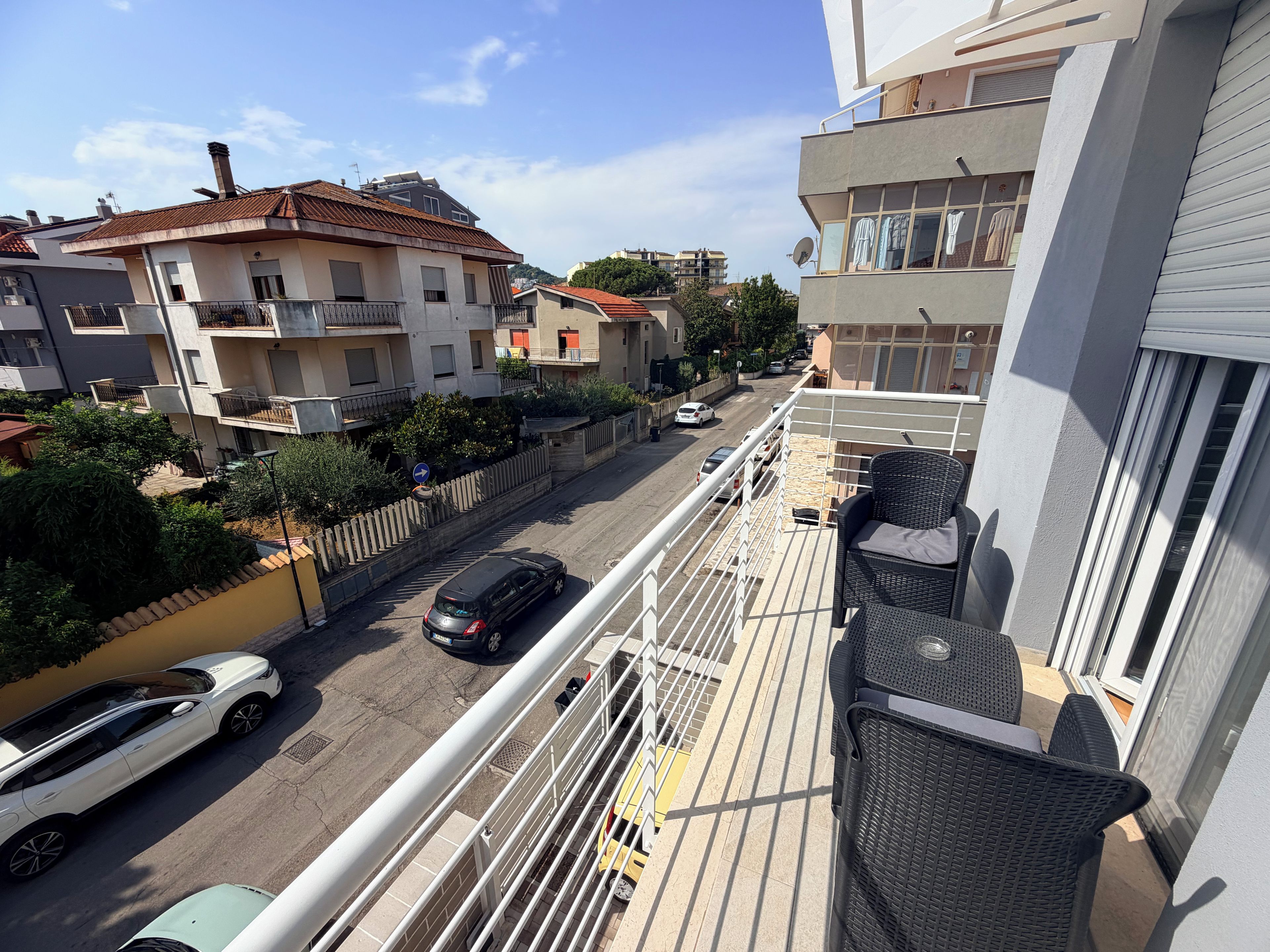 A balcony with outdoor furniture, a view of a street with parked cars, and residential buildings in the background.