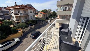 A balcony with outdoor furniture, a view of a street with parked cars, and residential buildings in the background.