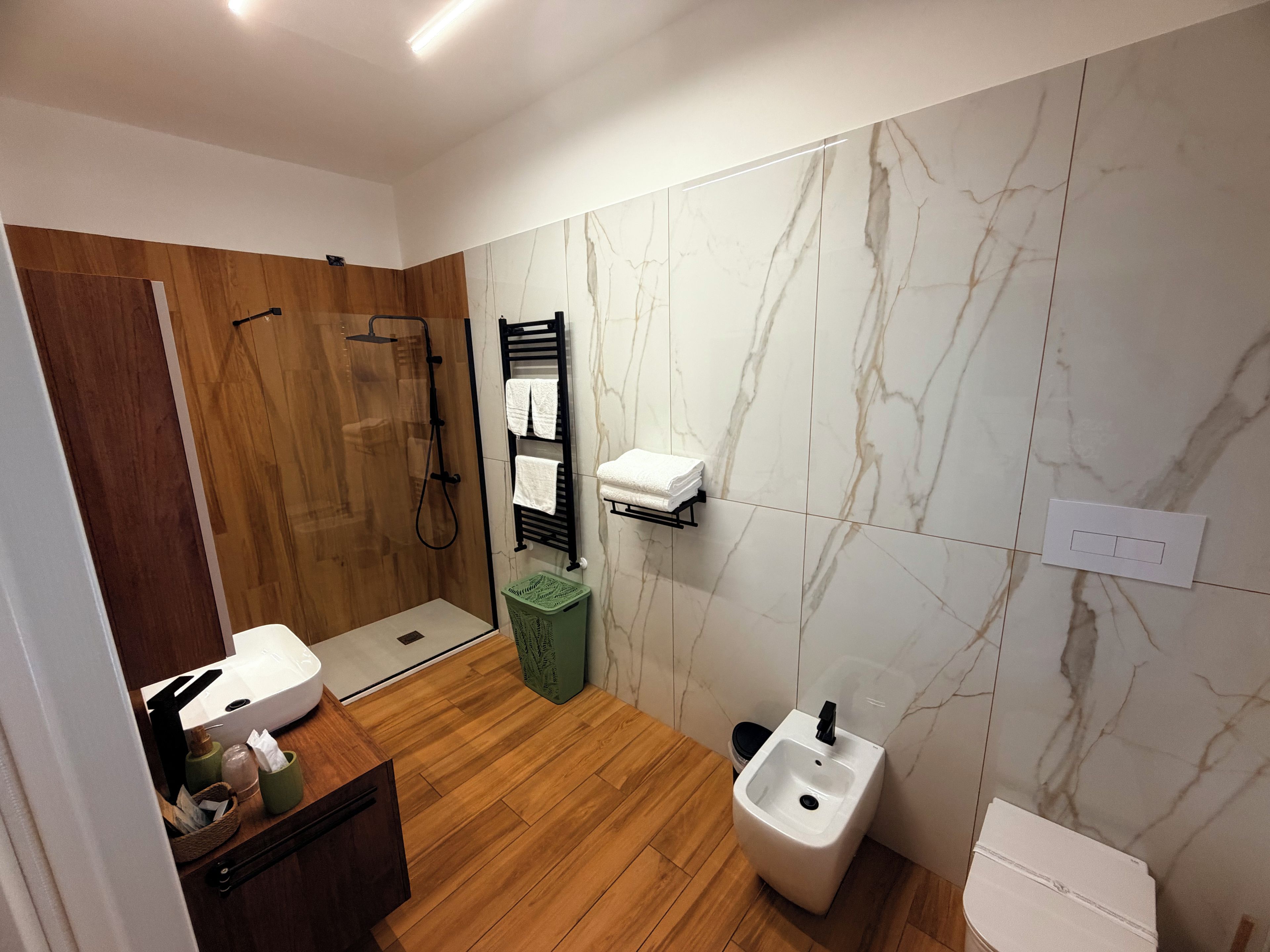 Ein modernes Badezimmer mit einer Duschkabine aus Holz, einem Waschbecken mit schwarzem Wasserhahn, einem Bidet und einer Wand mit Marmorplatten.