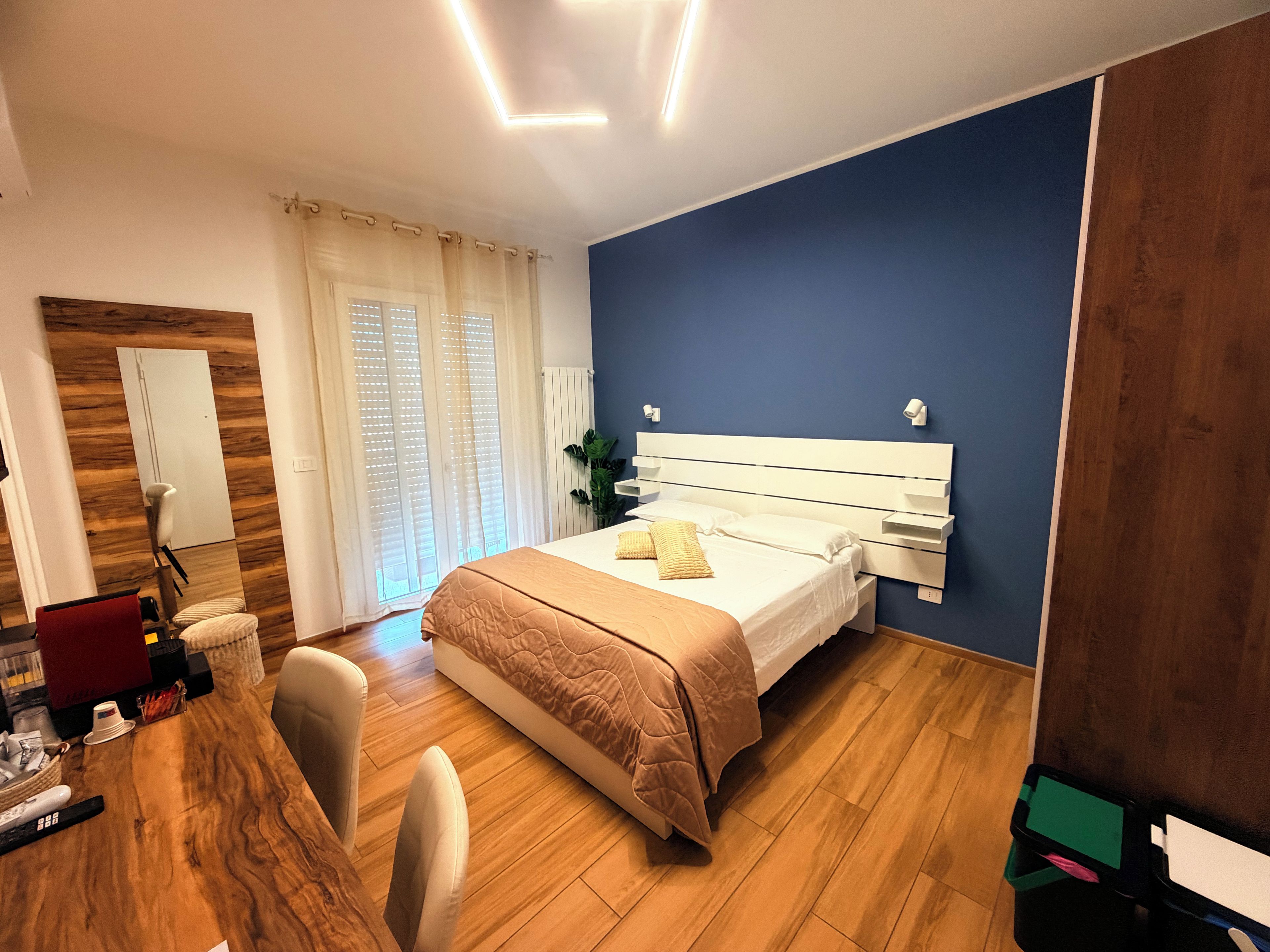 Habitación de hotel con piso de madera, una cama con cabecera blanca, un escritorio con silla y una ventana con cortinas.