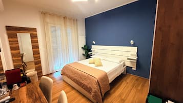 Habitación de hotel con piso de madera, una cama con cabecera blanca, un escritorio con silla y una ventana con cortinas.
