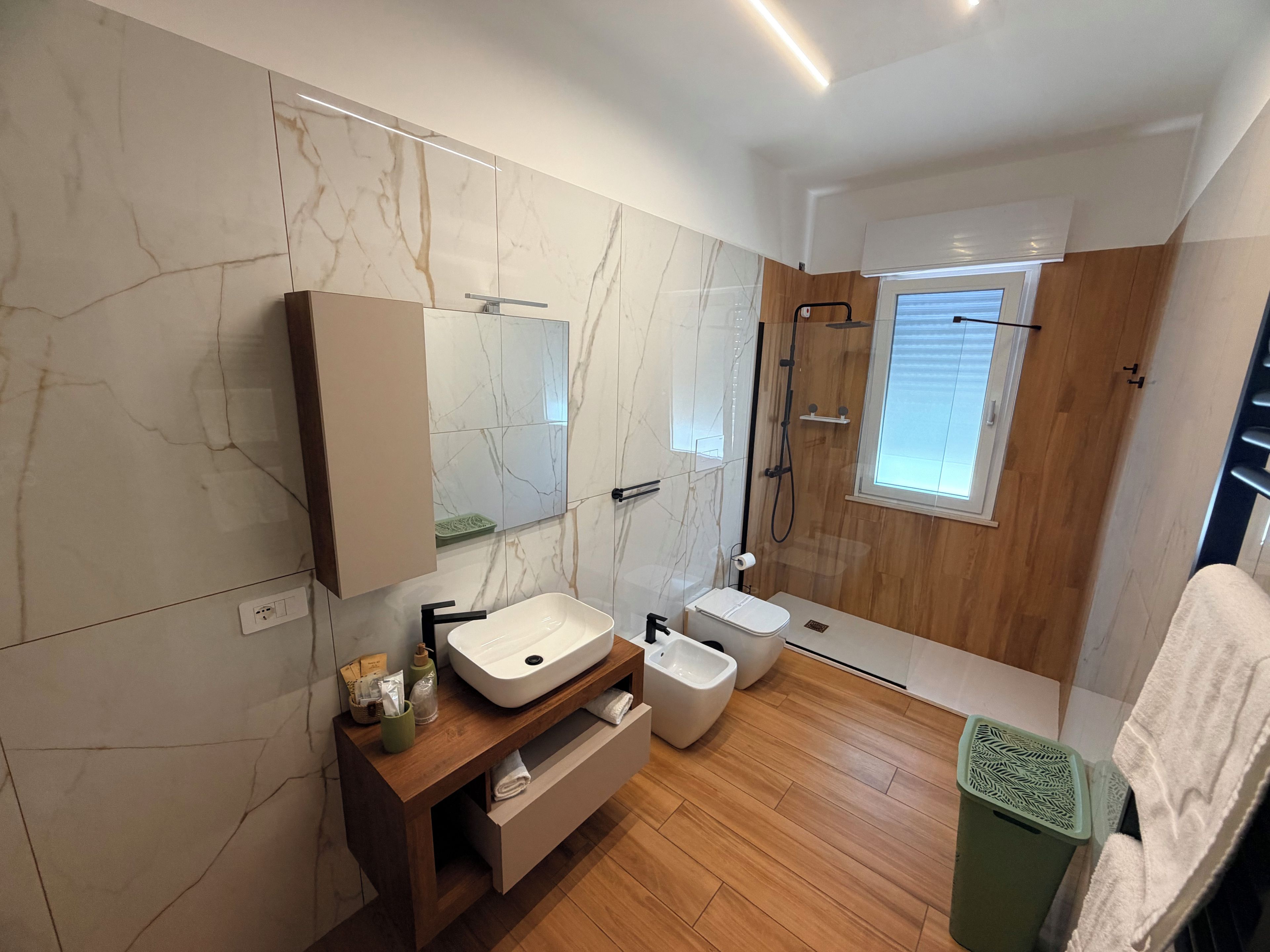 A modern bathroom with a marble wall, wooden vanity, a shower, and a window.