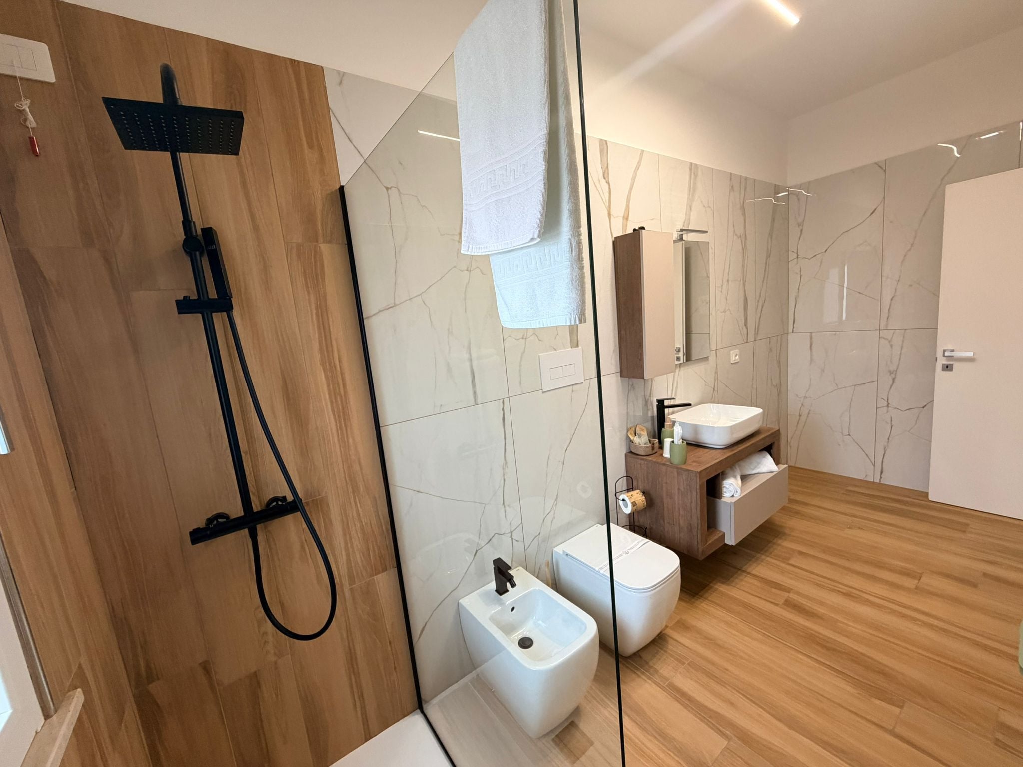 A modern bathroom with a wooden shower enclosure, a white sink, and a marble-tiled wall.