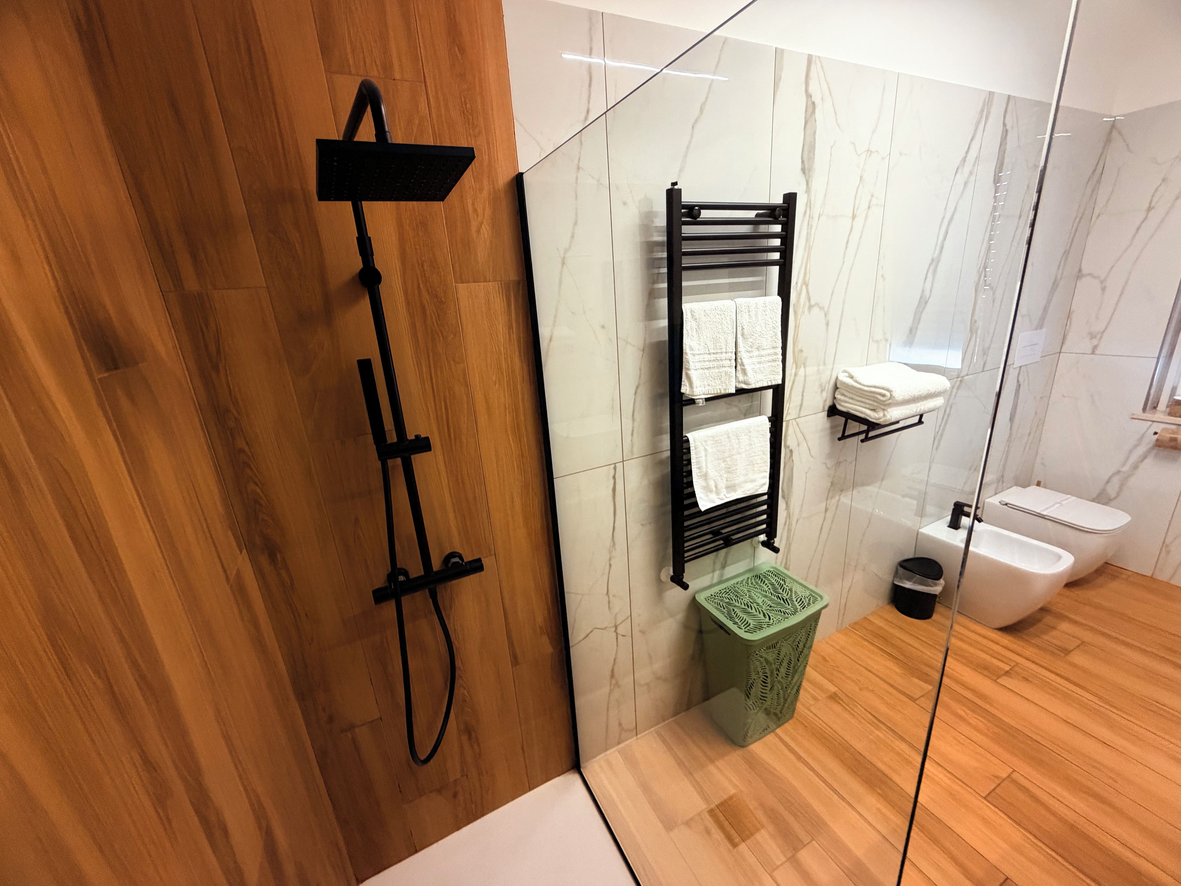 Une salle de bain moderne avec un plancher en bois, une pomme de douche noire, un porte-serviettes avec deux serviettes, une poubelle verte et des toilettes blanches.