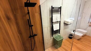 Une salle de bain moderne avec un plancher en bois, une pomme de douche noire, un porte-serviettes avec deux serviettes, une poubelle verte et des toilettes blanches.