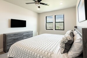 Un dormitorio con cama, televisor de pantalla plana en la pared, ventilador de techo y dos ventanas con vista al paisaje.