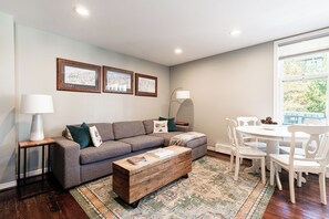 Ein modernes Wohnzimmer mit einem grauen Sofa, einem Holztisch, einer weißen Lampe und einem Esstisch mit Stühlen.