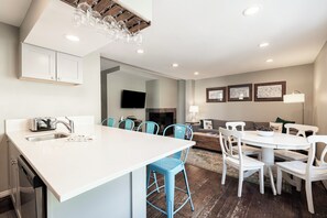 Eine moderne Küche mit Kochinsel, ein Essbereich mit rundem Tisch und Stühlen, ein Wohnzimmer mit Sofa und Kamin sowie ein an der Wand befestigter Flachbildfernseher.