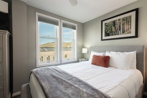 Habitación de hotel con una cama grande, una mesita de noche, una lámpara, un cuadro en la pared y vistas a un paisaje por las ventanas.