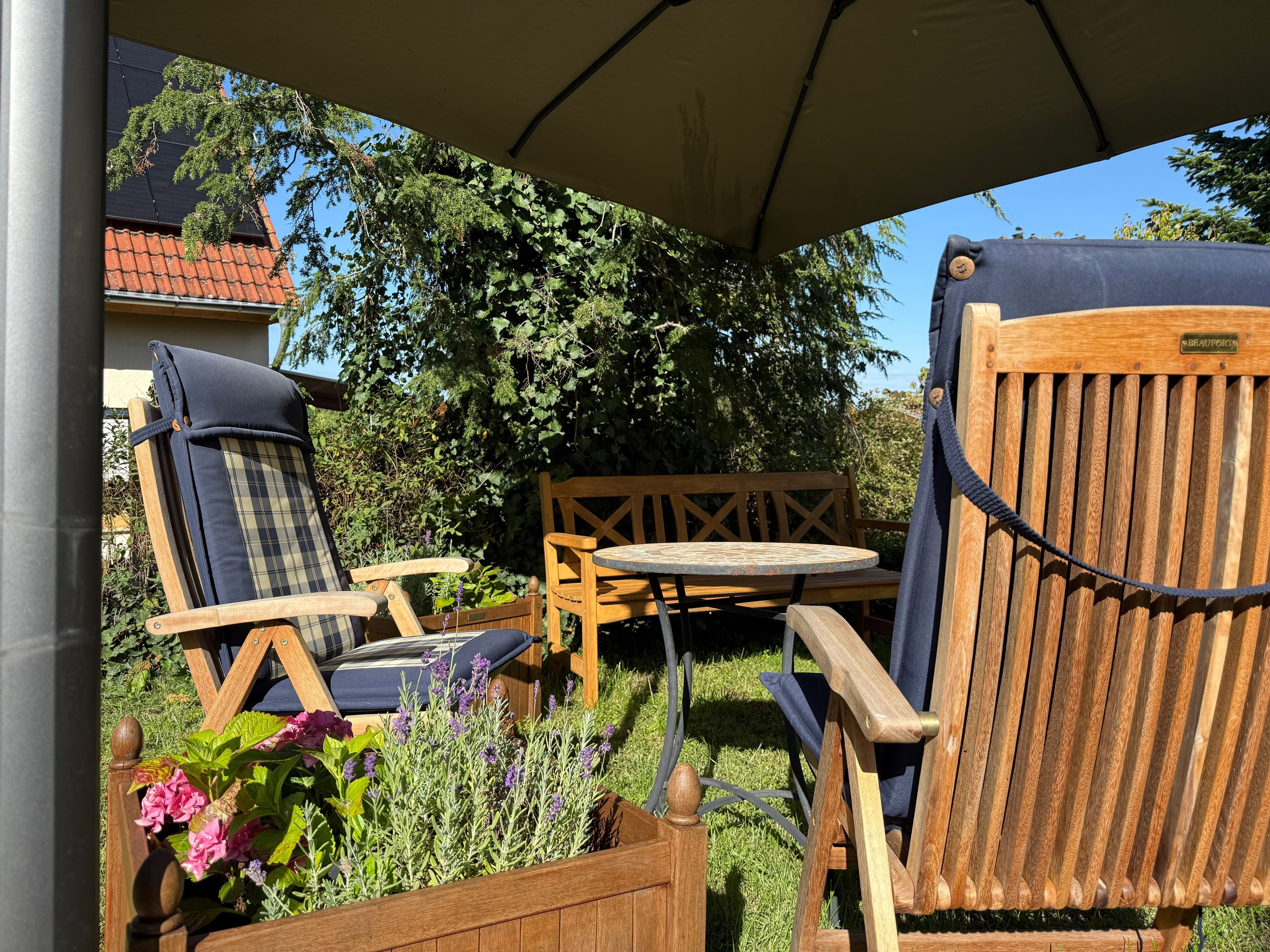 An outdoor seating area with wooden chairs and a table, surrounded by greenery and a building with a tiled roof.