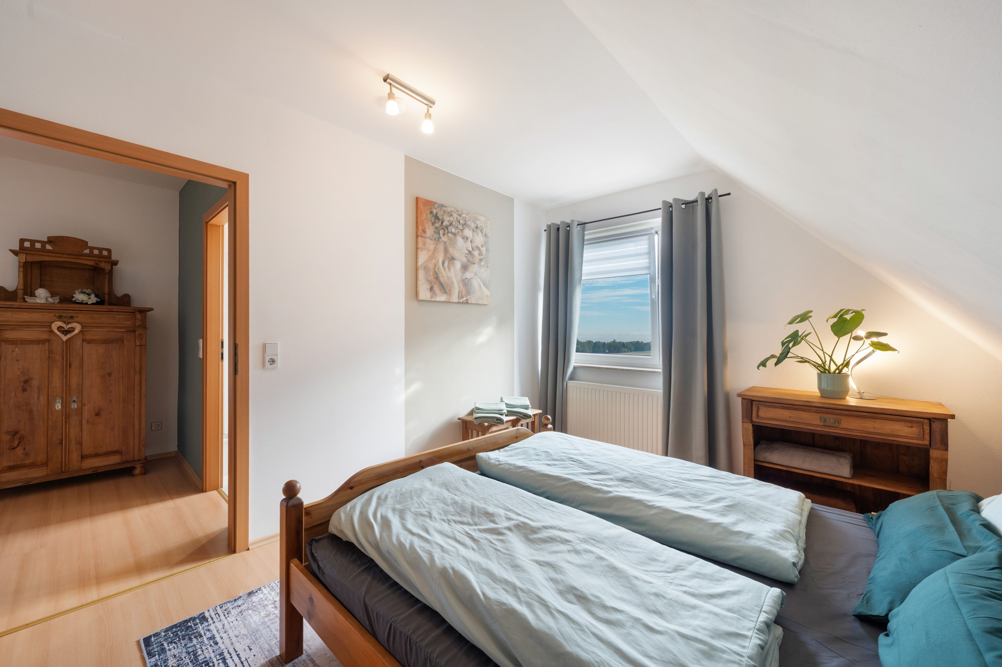 Une chambre avec deux lits, une armoire en bois, un tableau, une fenêtre avec des rideaux et une plante posée sur une table de chevet.
