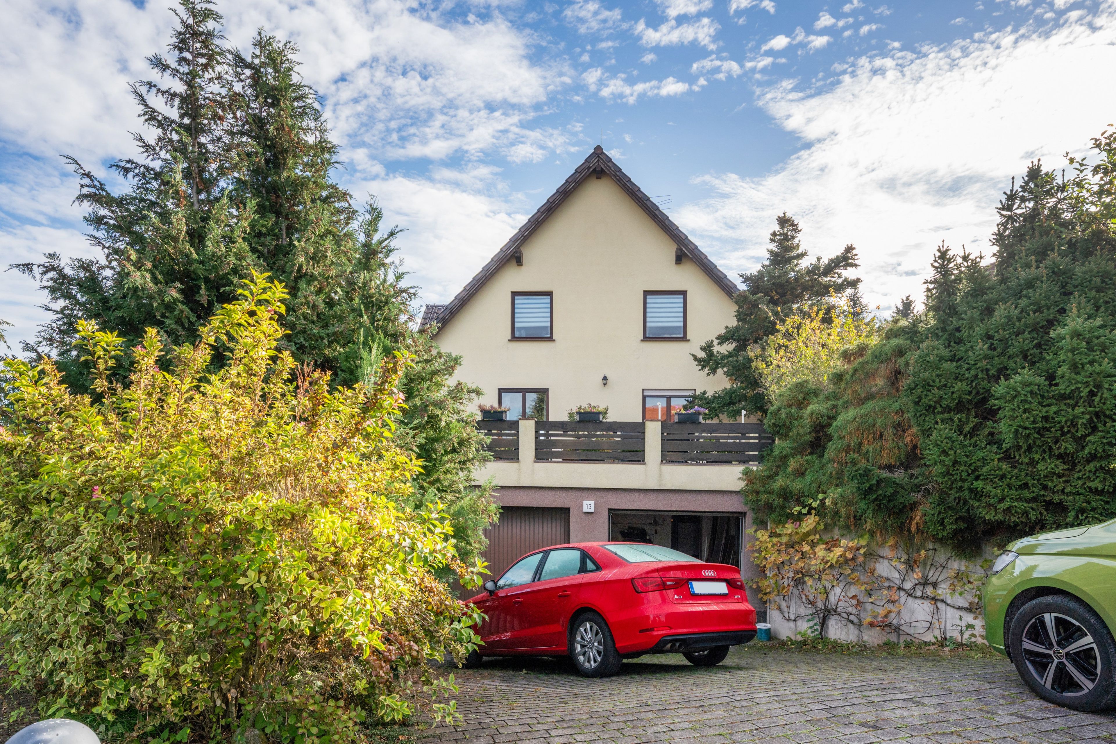 Une maison à deux étages avec un garage, entourée d’arbres et de voitures garées.