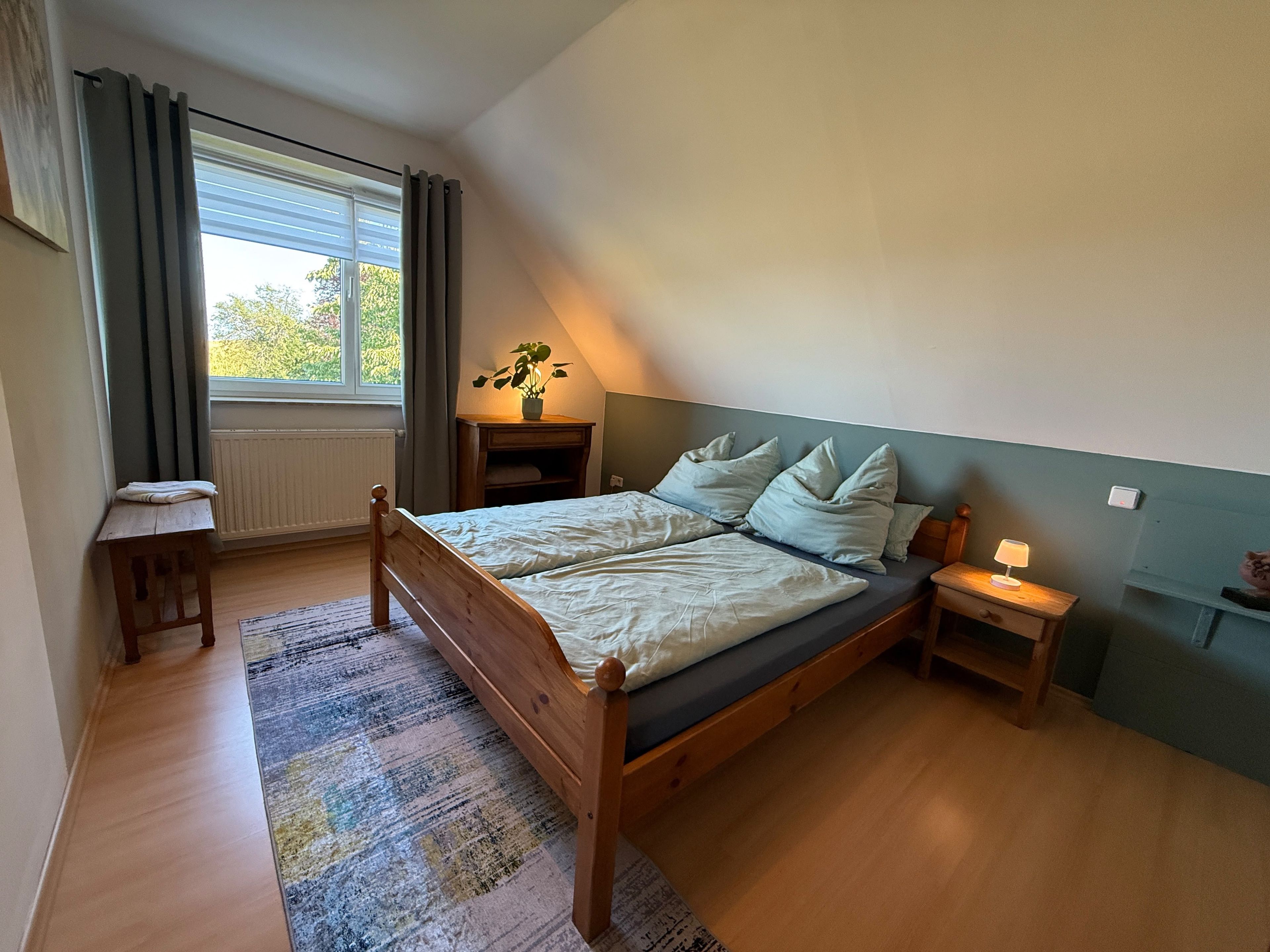 Een slaapkamer met een houten bed, een nachtkastje met een lamp, een klein tafeltje en een raam met gordijnen.