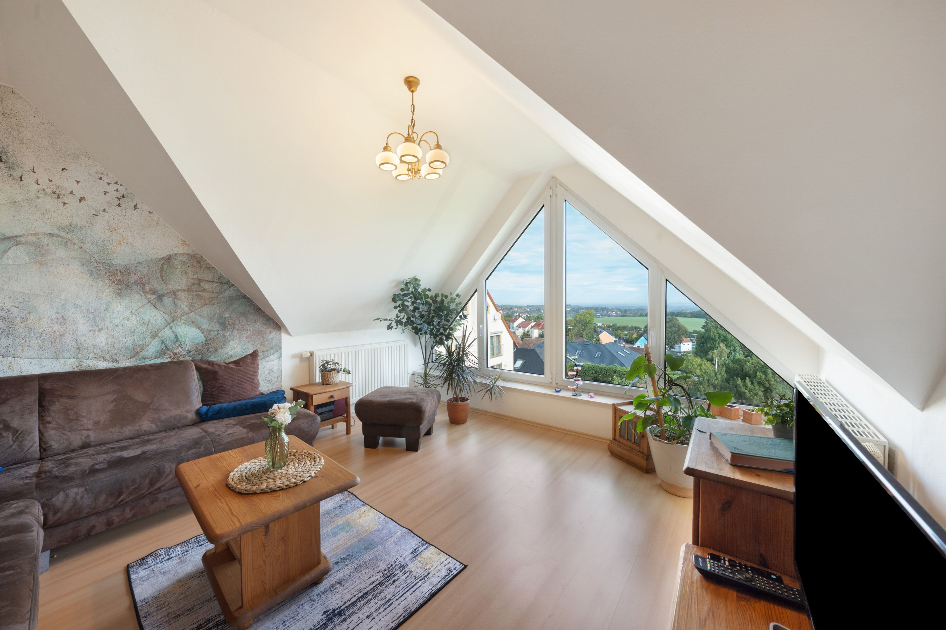 Eine moderne Loft-Wohnung mit schrägem Dach, einem großen Fenster mit Blick auf die Stadt, einer gemütlichen Sitzecke mit Sofa und Hocker sowie einem Holztisch.