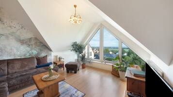 Eine moderne Loft-Wohnung mit schrägem Dach, einem großen Fenster mit Blick auf die Stadt, einer gemütlichen Sitzecke mit Sofa und Hocker sowie einem Holztisch.