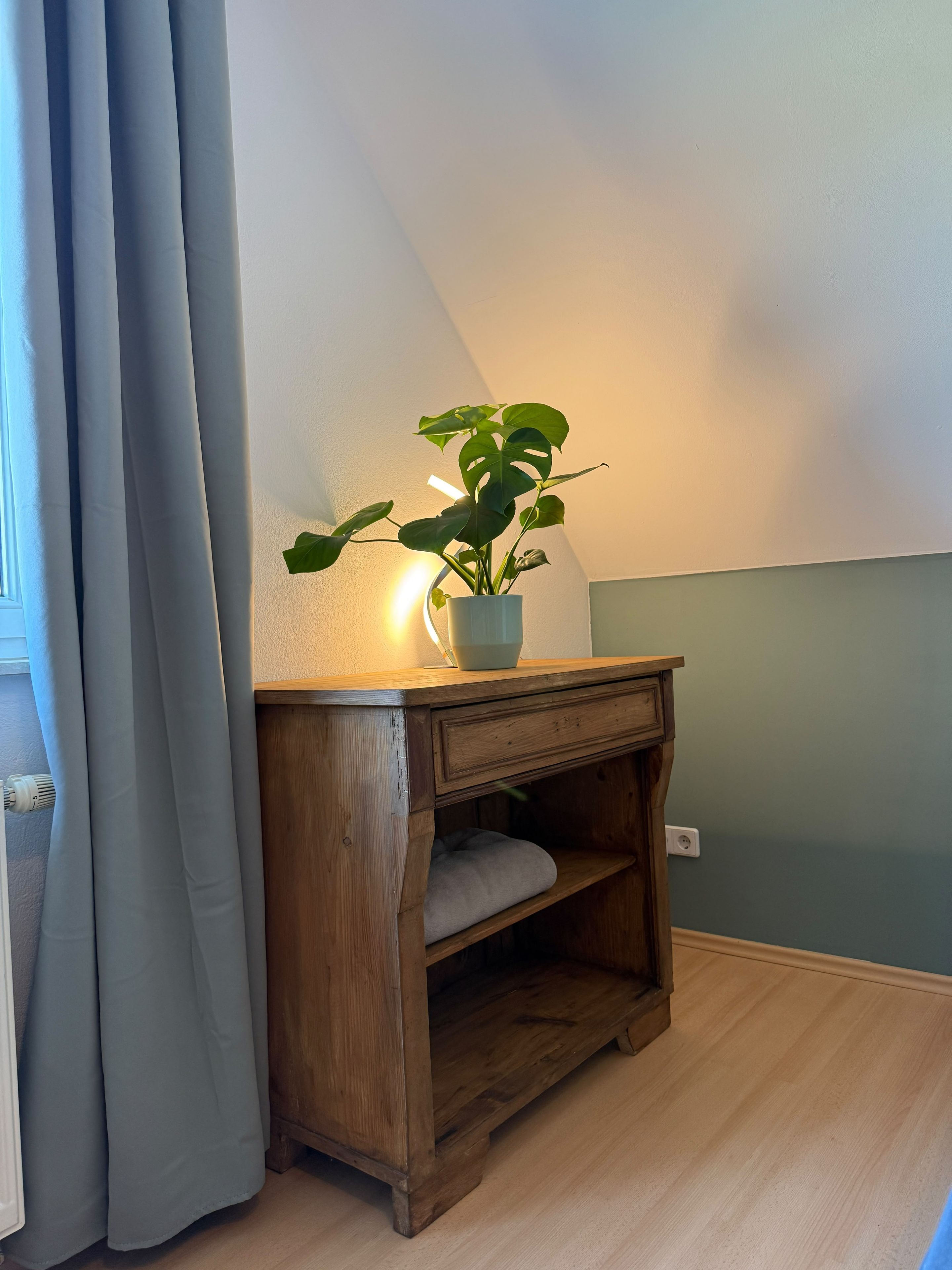 A corner of a room with a wooden nightstand, a potted plant, and a window with curtains.