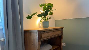A corner of a room with a wooden nightstand, a potted plant, and a window with curtains.
