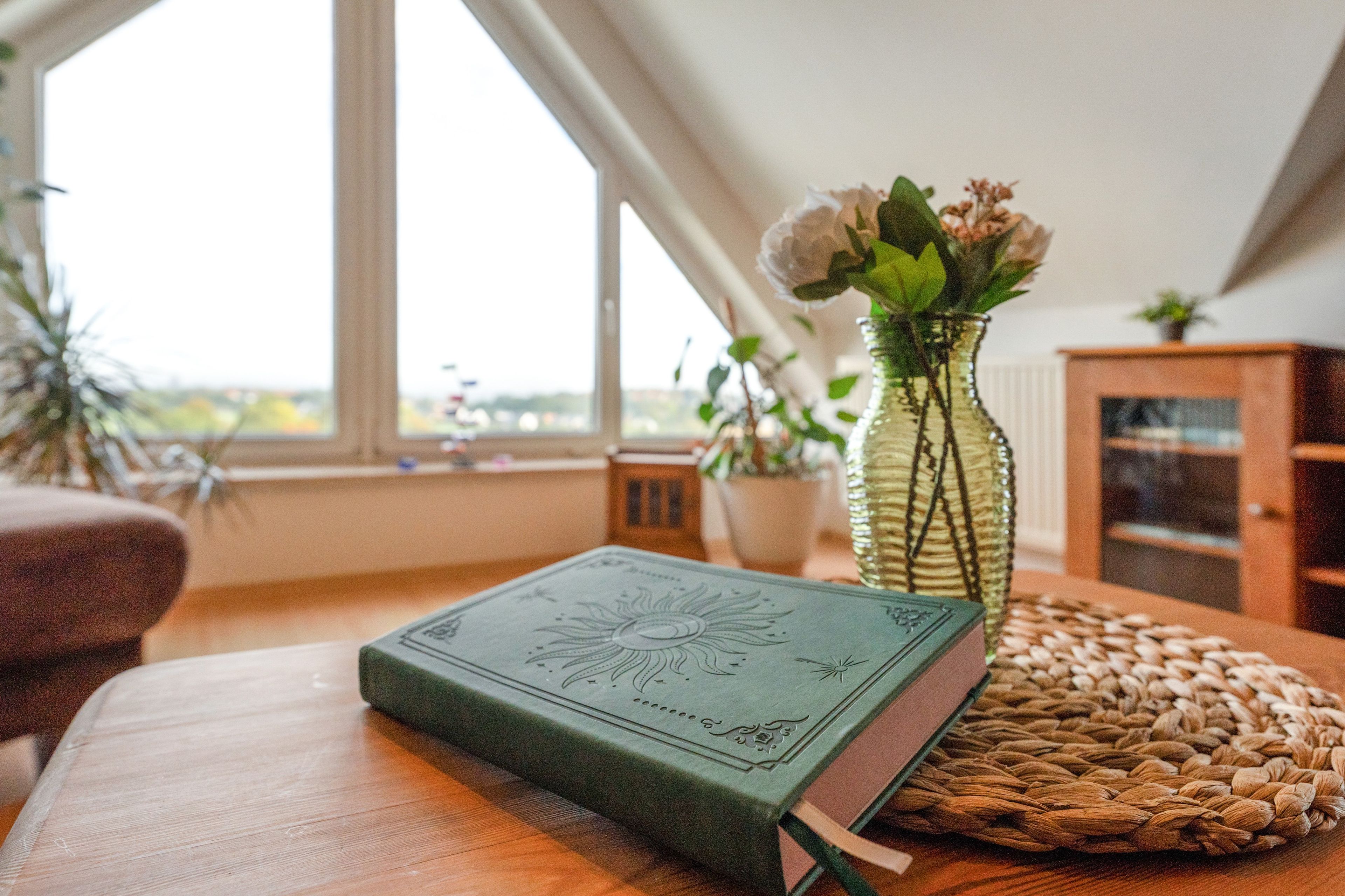 Ein Zimmer mit einem Holztisch, einem grünen Buch, einer Vase mit Blumen und Blick durch ein Fenster.
