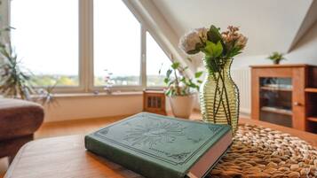 Ein Zimmer mit einem Holztisch, einem grünen Buch, einer Vase mit Blumen und Blick durch ein Fenster.