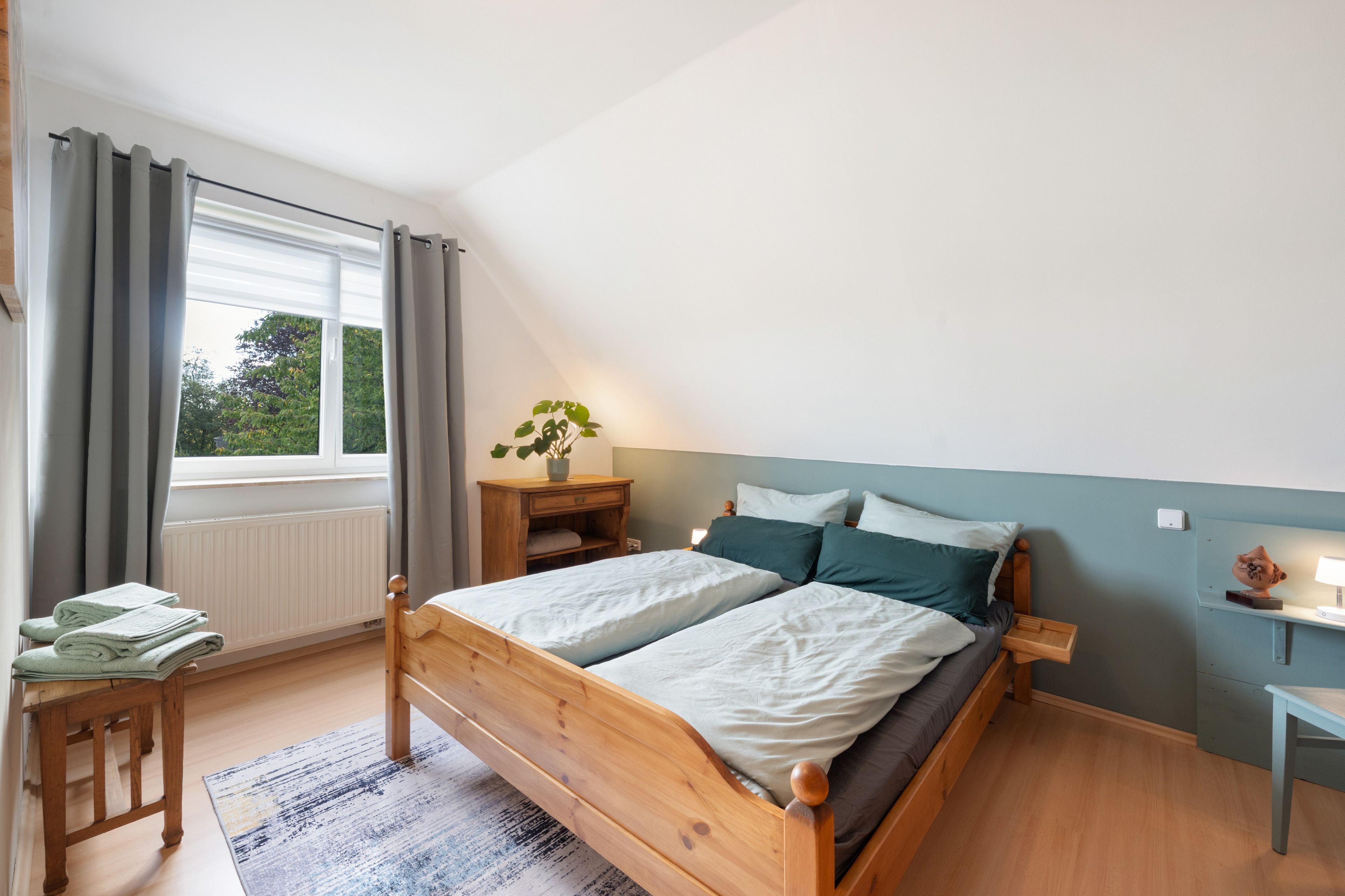 Ein Schlafzimmer mit einem hölzernen Bett, einem Nachttisch, einer Fensterbank und einem Vorhang vor dem Fenster.