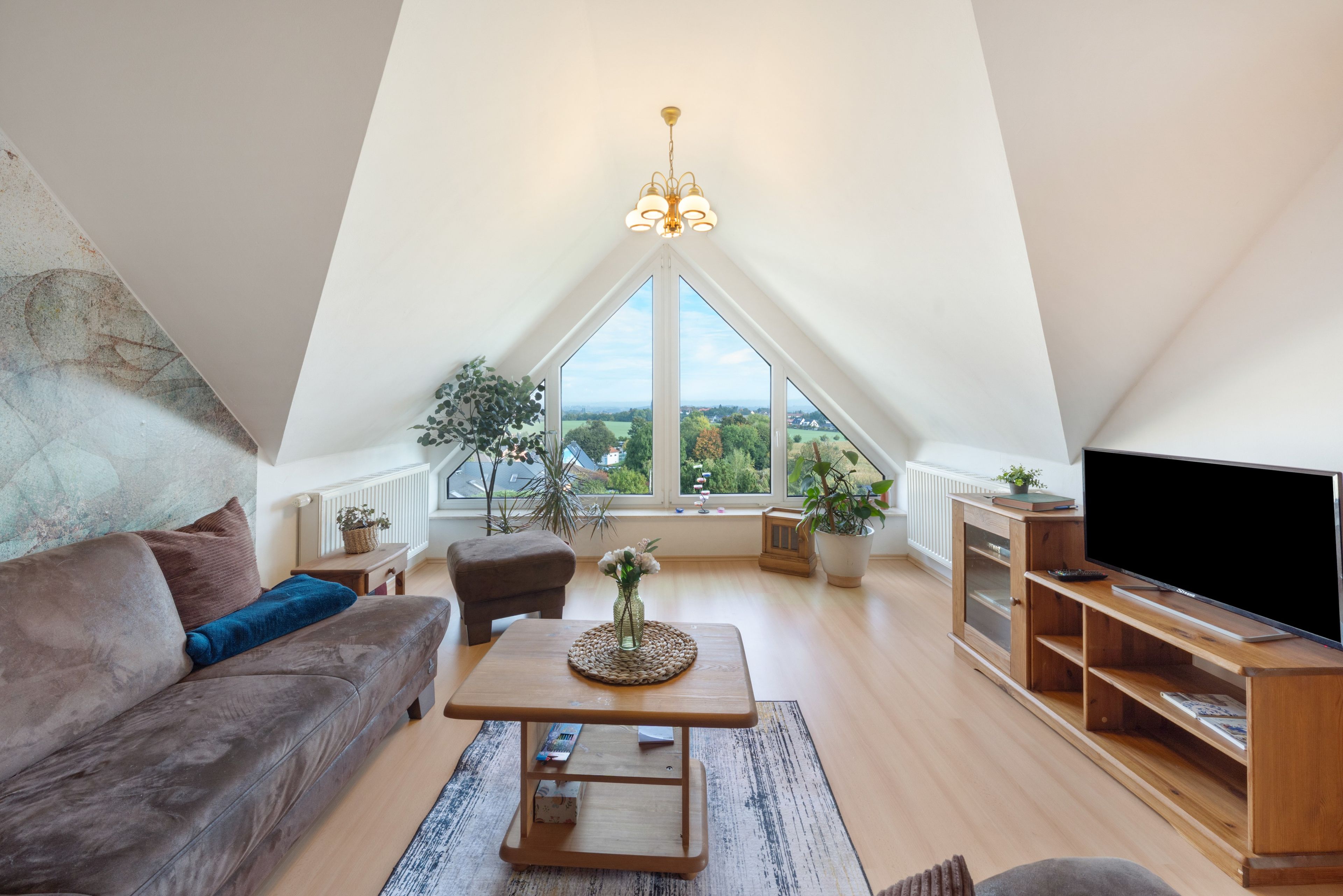 Geräumiges Loft mit hohen Decken, einem großen Fenster mit Blick auf die Landschaft, einer gemütlichen Sitzecke mit Sofa und Hocker, einem Holztisch und einem Fernseher auf einem Holzgestell.