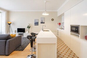 A modern kitchen with a central island, a dining area with a grey sofa and black chairs, and a flat-screen TV. - Virginie Almada Apartment (Porto)