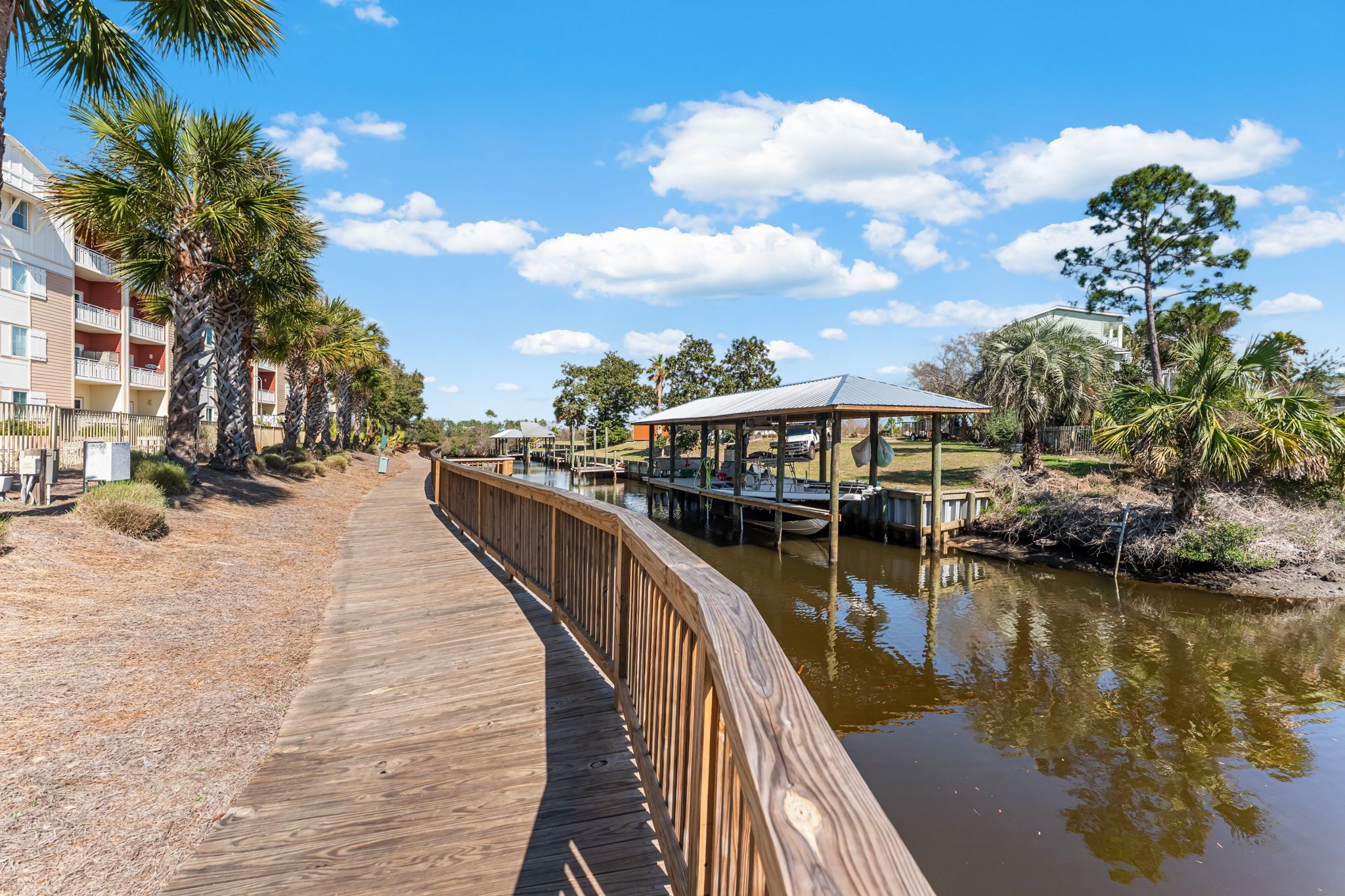 A wooden boardwalk leading to a dock area with a covered structure, surrounded by palm trees and a clear sky.