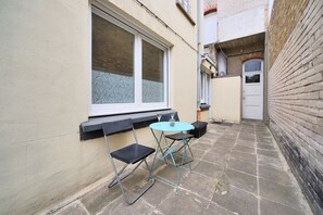 A small outdoor seating area with a table and two chairs, a window with a view, and a door to a brick building. - La Maison Petite Pause - plage à 150 m (Bray-Dunes)