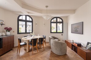Ein Essbereich mit einem runden Tisch, gedeckt für vier Personen, umgeben von Holzstühlen, einem grauen Sessel und einer Holzkommode mit Wasserkocher und Blumen.