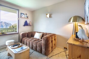 A cozy room with a plush brown sofa, a small wooden side table with a lamp, and a view of mountains through a window. - L’écrin 206 - Quartier des Bergers - Proximité immédiate ESF (Huez)