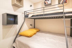 Ein Etagenbett mit weißer Matratze und gelbem Kopfkissen, ein kleines Regal mit Büchern und ein an der Wand befestigter Fernseher.