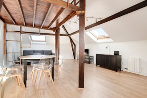A spacious loft with a high ceiling, exposed wooden beams, a skylight, a round dining table with chairs, a sofa, and a dark wooden cabinet. - Le Vivaldi - Proche de la Gare (Nancy)