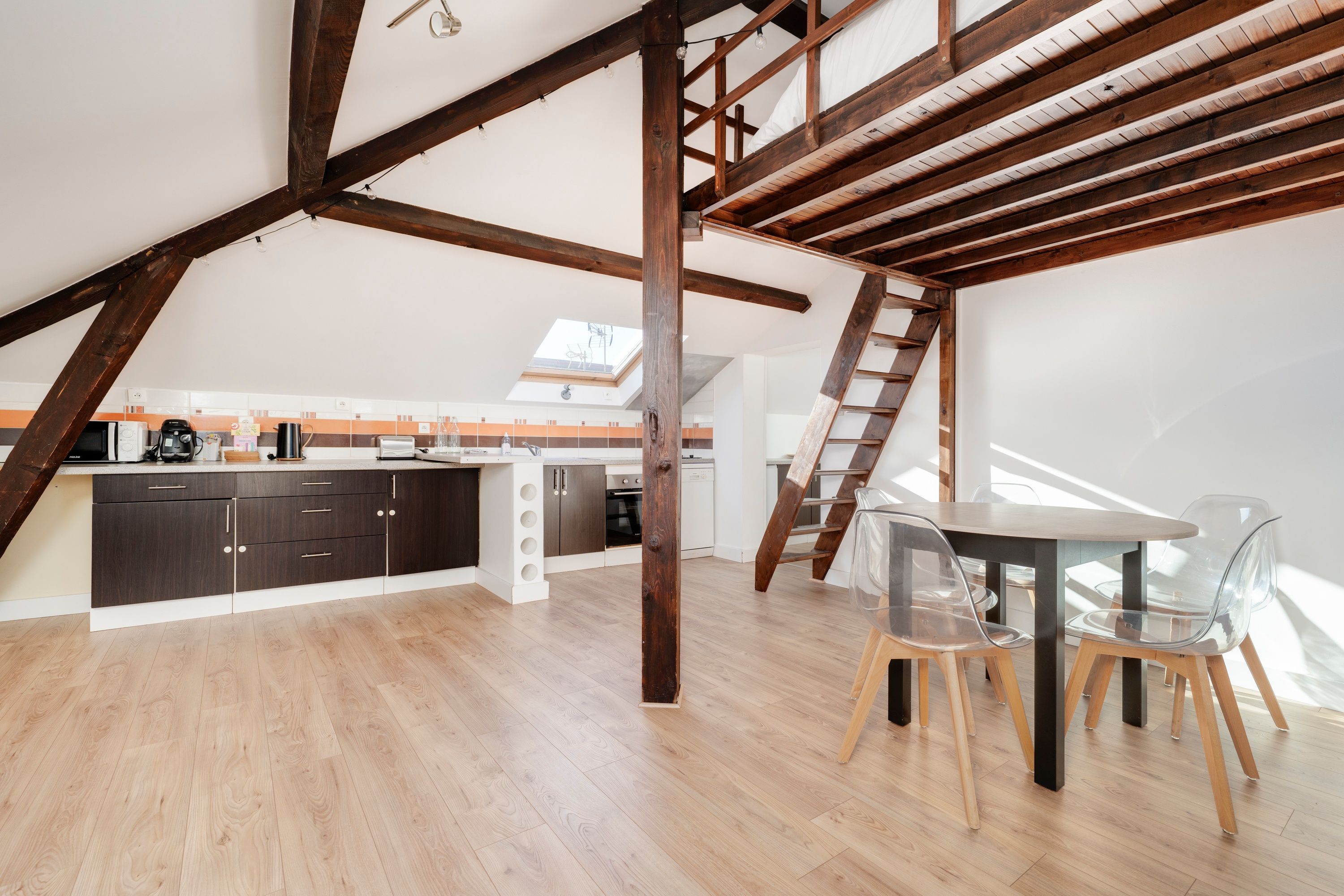 Un loft moderne avec des poutres apparentes, une cuisine dotée de meubles sombres et un coin repas avec des chaises transparentes.
