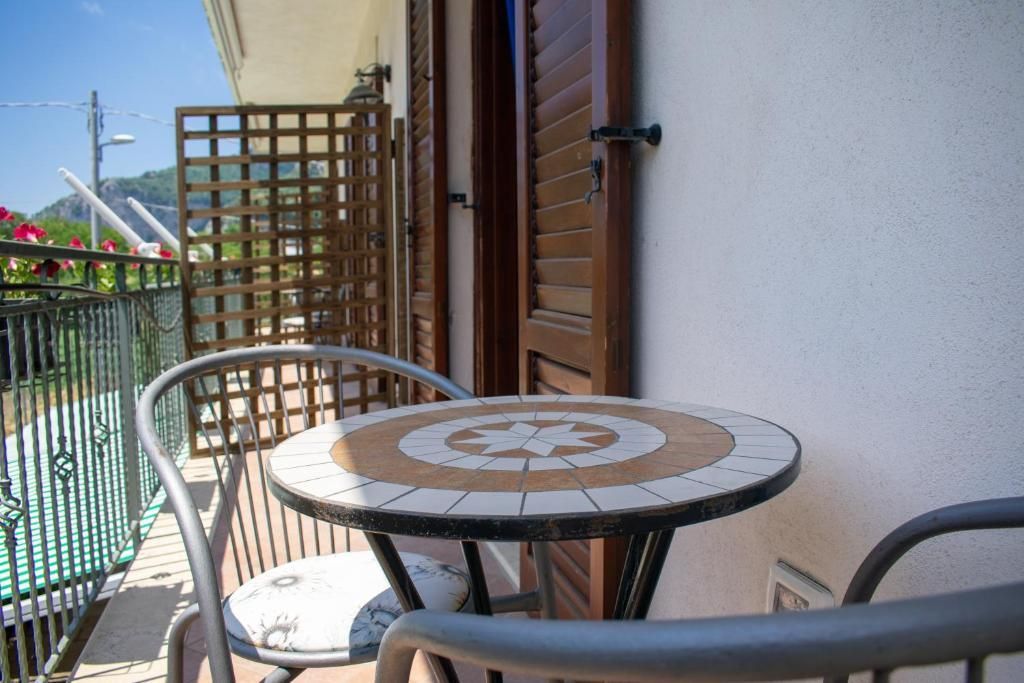 Une petite table d’extérieur, recouverte de carreaux, entourée de chaises en métal, placée contre un mur d’immeuble orné de volets en bois.