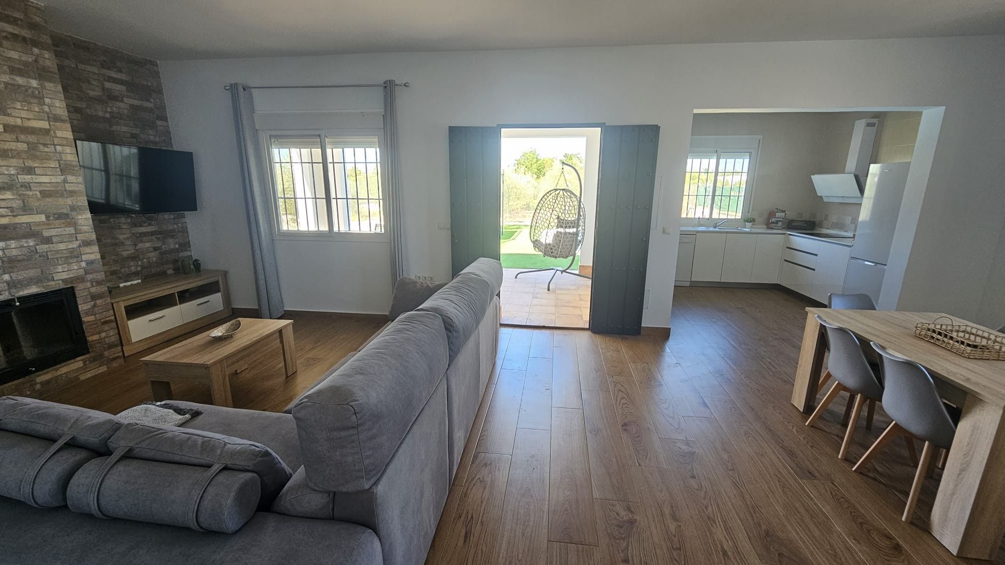 Una sala moderna con un sofá gris, una mesa de centro de madera y un televisor montado en una pared de ladrillo. Hay una zona de cocina con armarios blancos y una mesa de comedor con sillas.