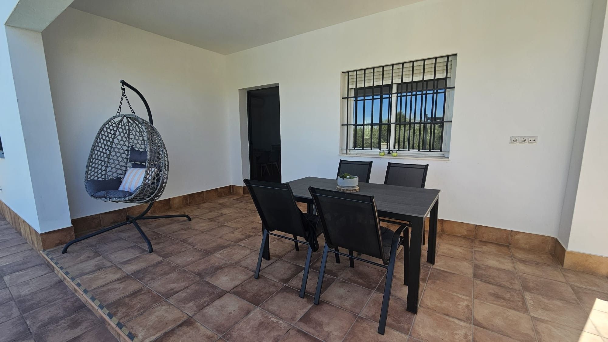 Un espace repas comprenant une table, des chaises, un fauteuil suspendu et une fenêtre avec des barreaux.
