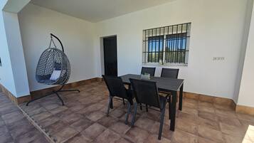 Un espace repas comprenant une table, des chaises, un fauteuil suspendu et une fenêtre avec des barreaux.