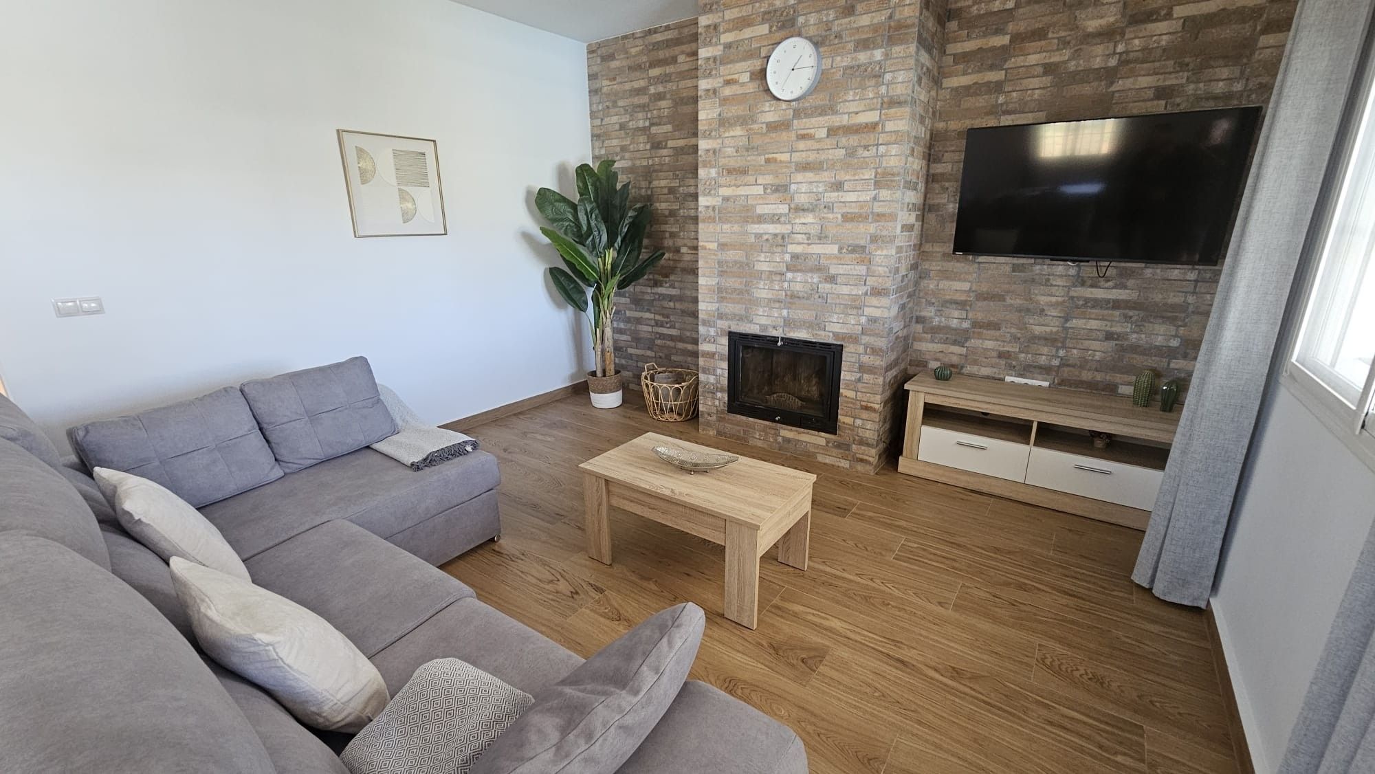 Una sala de estar con un sofá gris, una mesa de centro de madera, una chimenea de ladrillo, un televisor de pantalla plana y una planta en maceta.