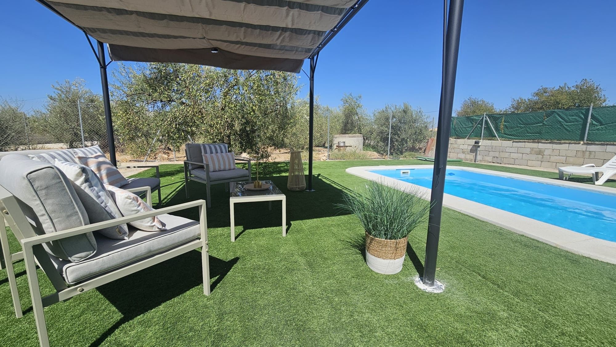 Patio con césped artificial, piscina, zona de estar techada y mesa.