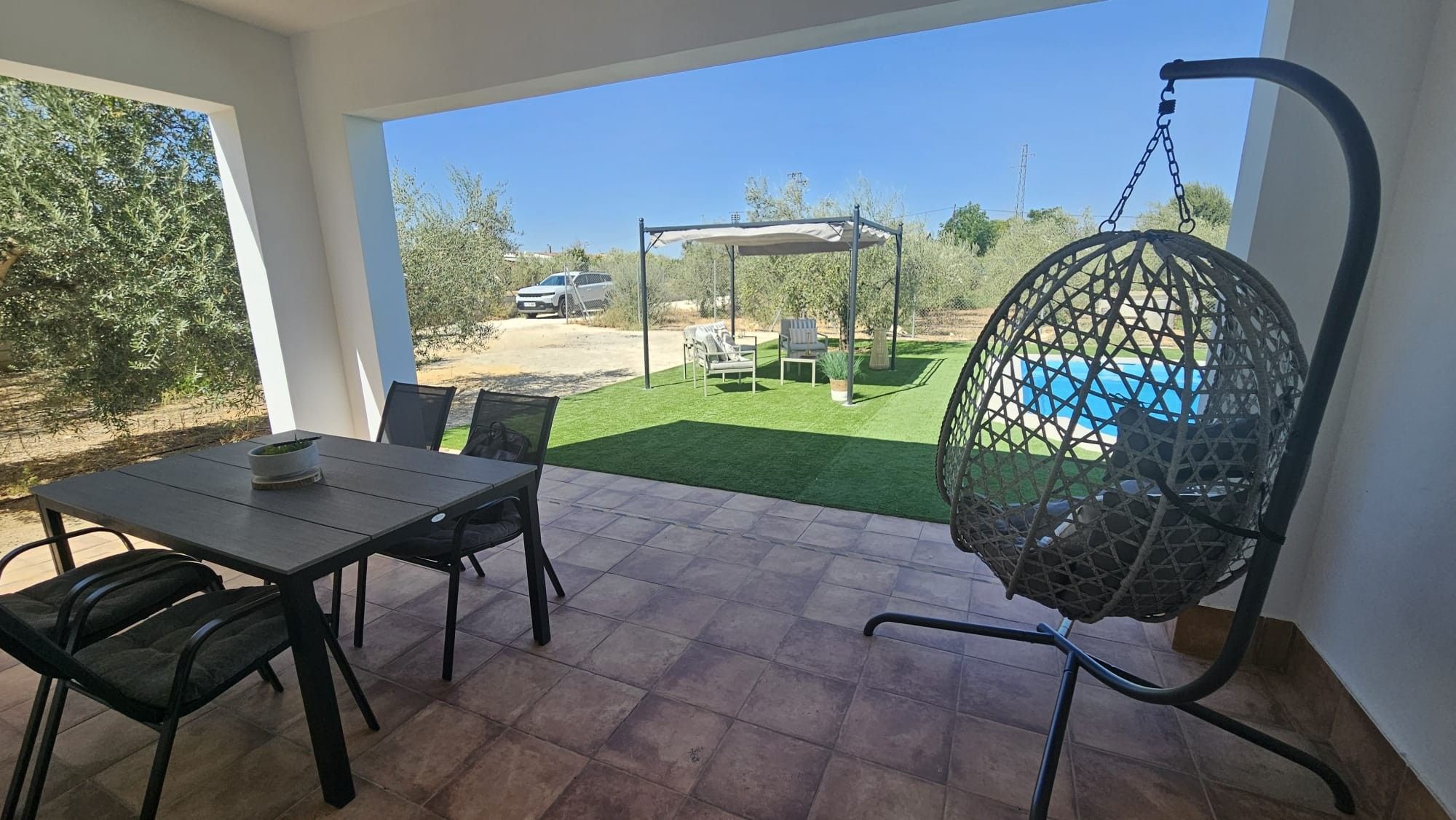 A patio with a table and chairs, a hanging chair, and a view of a pool and garden.