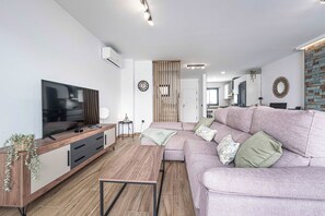 Uma sala de estar moderna com uma televisão de tela plana, um móvel de entretenimento de madeira, um sofá grande com almofadas e uma área de jantar ao fundo.