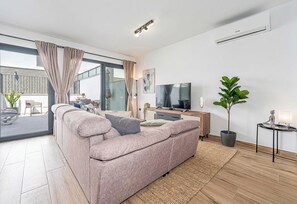 Un salon moderne avec un grand canapé, un téléviseur à écran plat, une plante en pot et un balcon agrémenté de rideaux.