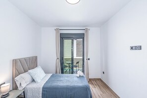 Quarto com cama, mesa de cabeceira, abajur, janela com cortinas e vista para o exterior.