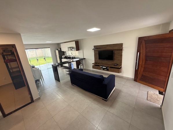 A modern living room with a sofa, a TV mounted on a wooden wall, and a kitchen area with a fridge and sink.