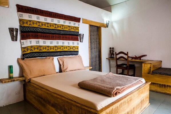 A bed with a wooden frame and a headboard with patterned fabric. There is a bedside table with a lamp and a small wooden stool.
